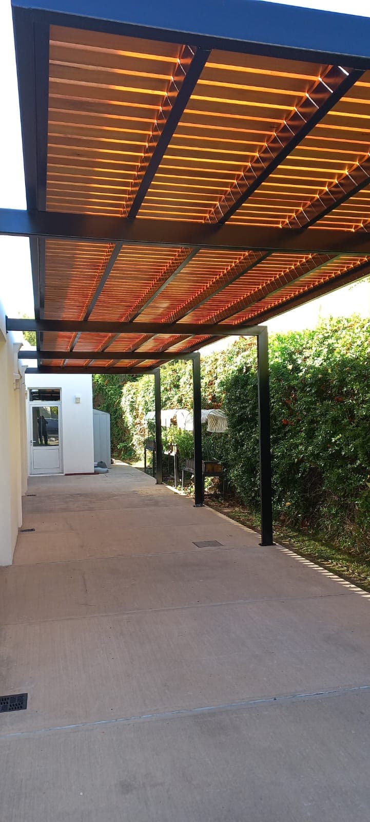 Pérgola metálica con listones de madera sobre un acceso lateral con cerco verde